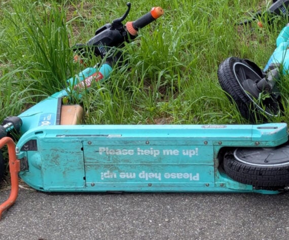 A turquoise electric scooter lies on its side in the grass next to a paved path. The side of the scooter reads, &ldquo;Please help me up!&rdquo; in white text, printed both right-side up and upside down.