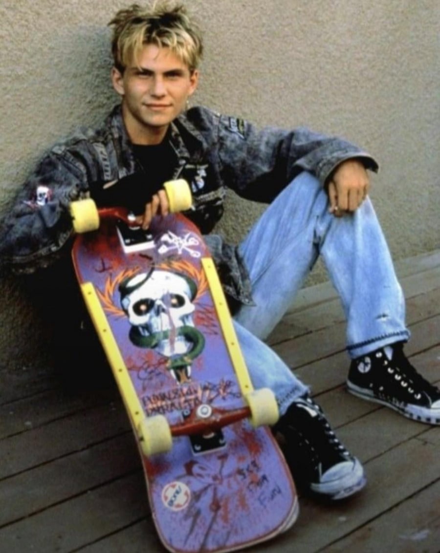 A young person with blond hair in a denim jacket and jeans sits on a wooden floor, holding a skateboard with colorful artwork and yellow wheels. The person is wearing black Converse sneakers and is leaning against a plain wall.