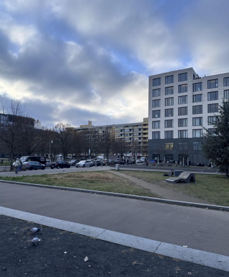 A park with bare trees, a paved walkway, and grass patches is shown. In the background are modern, mid-rise apartment buildings. The sky is mostly cloudy, and a few pigeons are visible in the foreground.