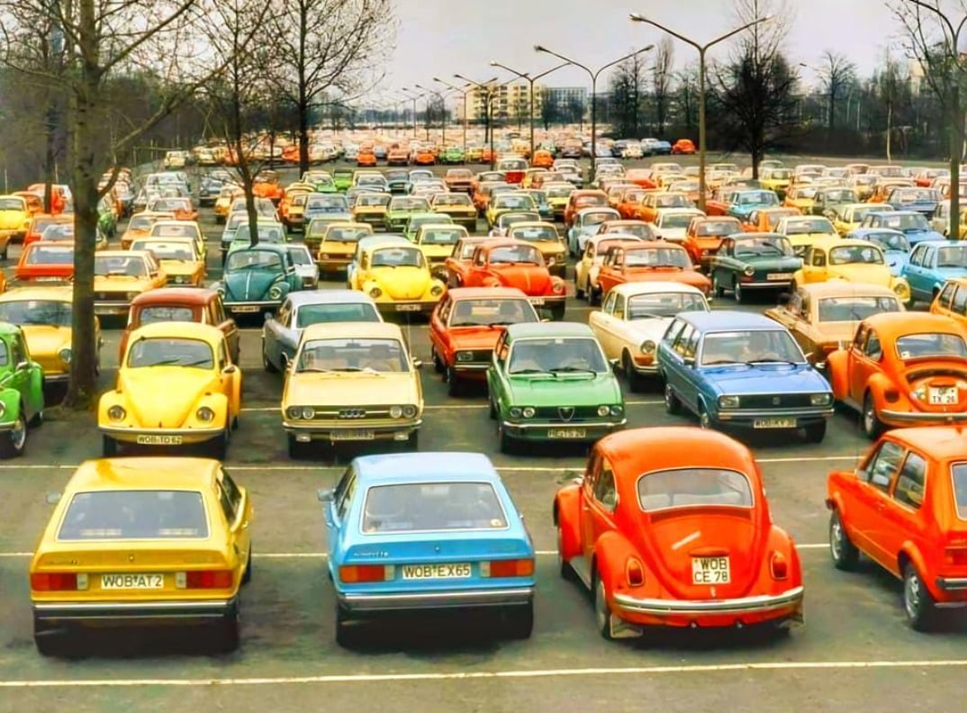 A large parking lot filled with colorful vintage cars from the 1970s, including several Volkswagen Beetles, with leafless trees and apartment buildings visible in the background.