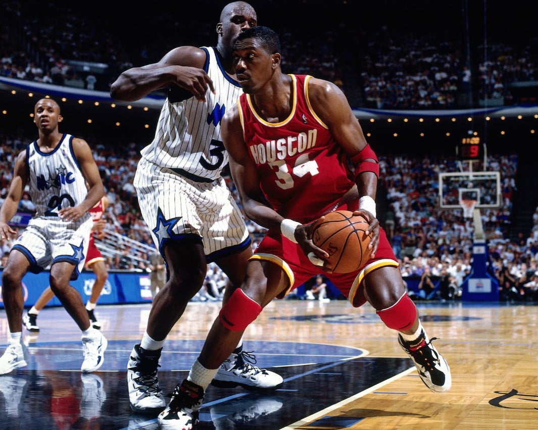 A basketball player wearing a red Houston Rockets jersey drives to the basket while being closely guarded by a player in a white Orlando Magic uniform during an NBA game in a packed arena.