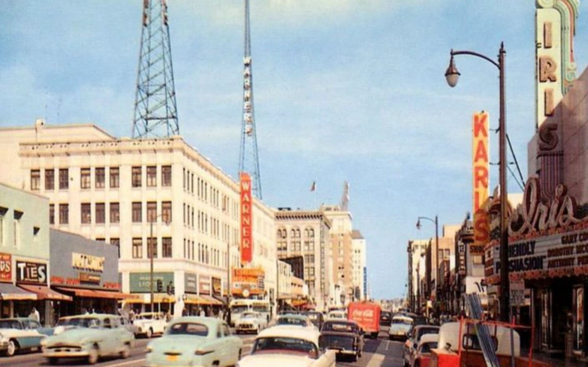 A busy 1950s city street lined with vintage cars, neon signs, and storefronts. People walk on sidewalks near shops and theaters. Two tall radio towers rise above the buildings under a blue sky.