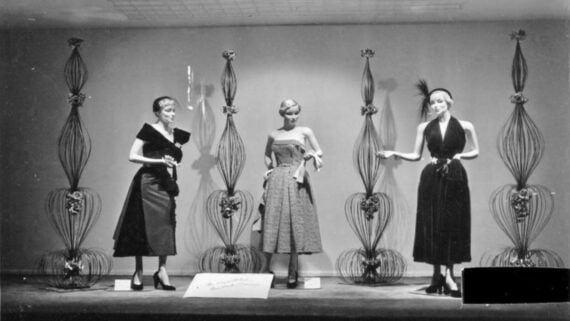 Three women in elegant 1950s-style dresses pose on a stage with decorative wire sculptures behind them. Each holds gloves or accessories, standing in dramatic, fashion-model poses under soft lighting.
