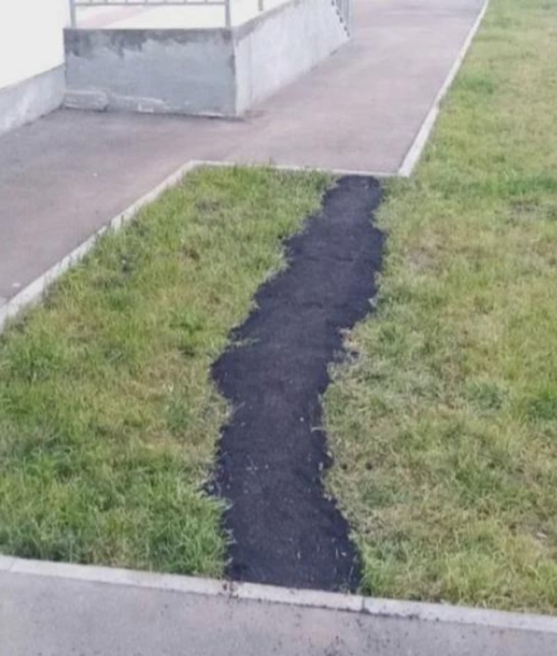 A grassy area with a narrow, uneven strip of black asphalt or pavement cutting through the grass, suggesting a poorly made path between two concrete sidewalks.