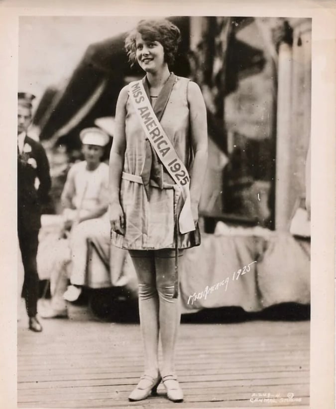 A young woman wearing a "Miss America 1925" sash stands and smiles outdoors, dressed in a sleeveless dress and tights. Two people, including a man in a sailor uniform, are seated in the blurred background.