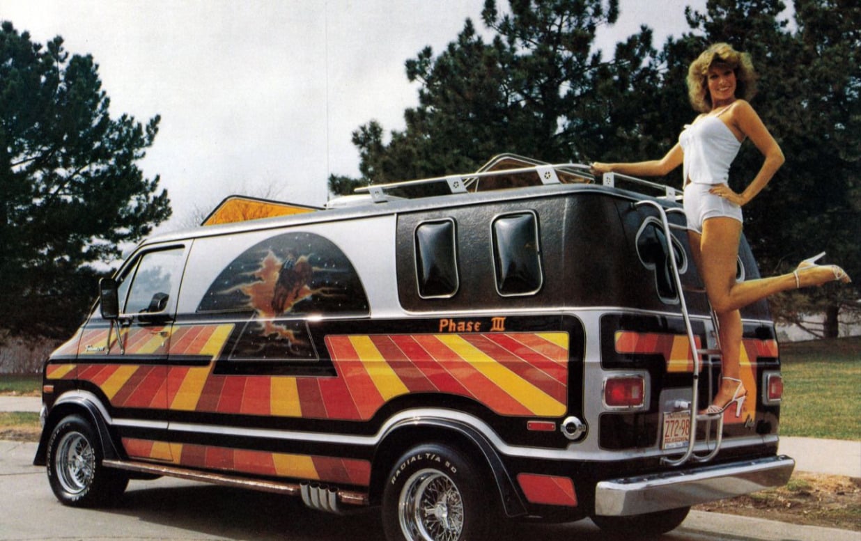 A woman in a white outfit poses with one leg raised on the rear bumper of a custom van featuring bright red and yellow stripes, chrome wheels, and an airbrushed mural on the side. Trees and grass are in the background.