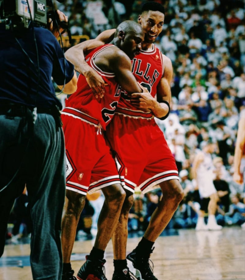 Two Chicago Bulls basketball players in red uniforms embrace on the court; one supports the other, who appears exhausted. A cameraman films them as a crowd watches in the background.