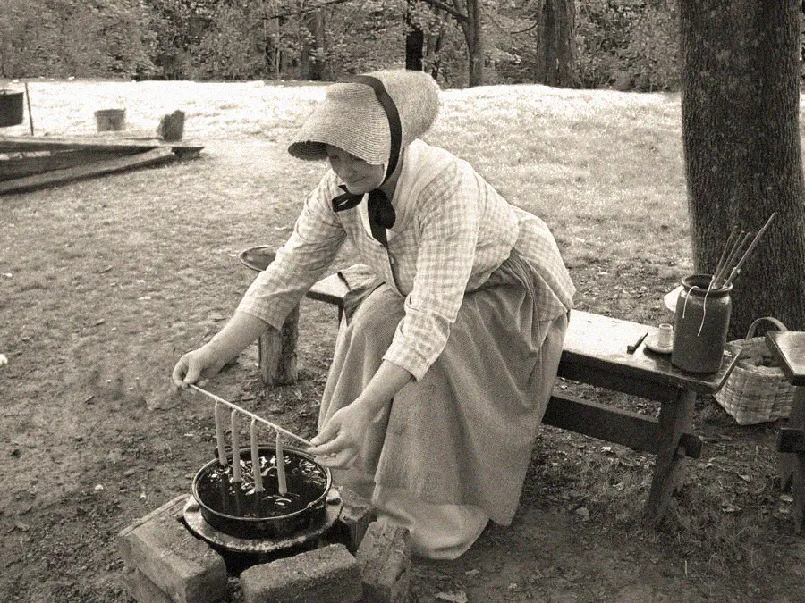 A woman in historical clothing and bonnet sits outdoors, dipping candles by hand into a pot over a fire pit, with a wooden bench and trees in the background. The photo is in black and white.
