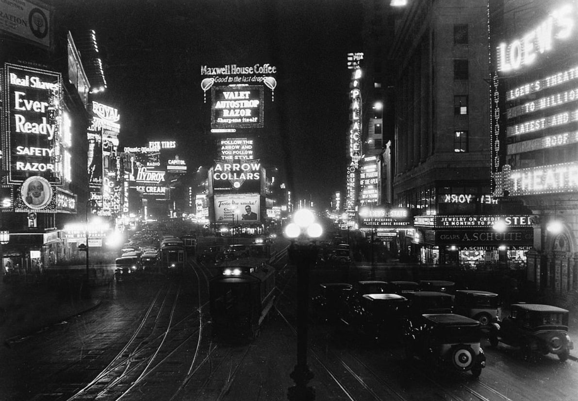 A black-and-white photo of Times Square at night in the 1920s, with vintage cars, streetcars, and bright neon signs advertising coffee, theaters, and razors, illuminating the bustling city street.