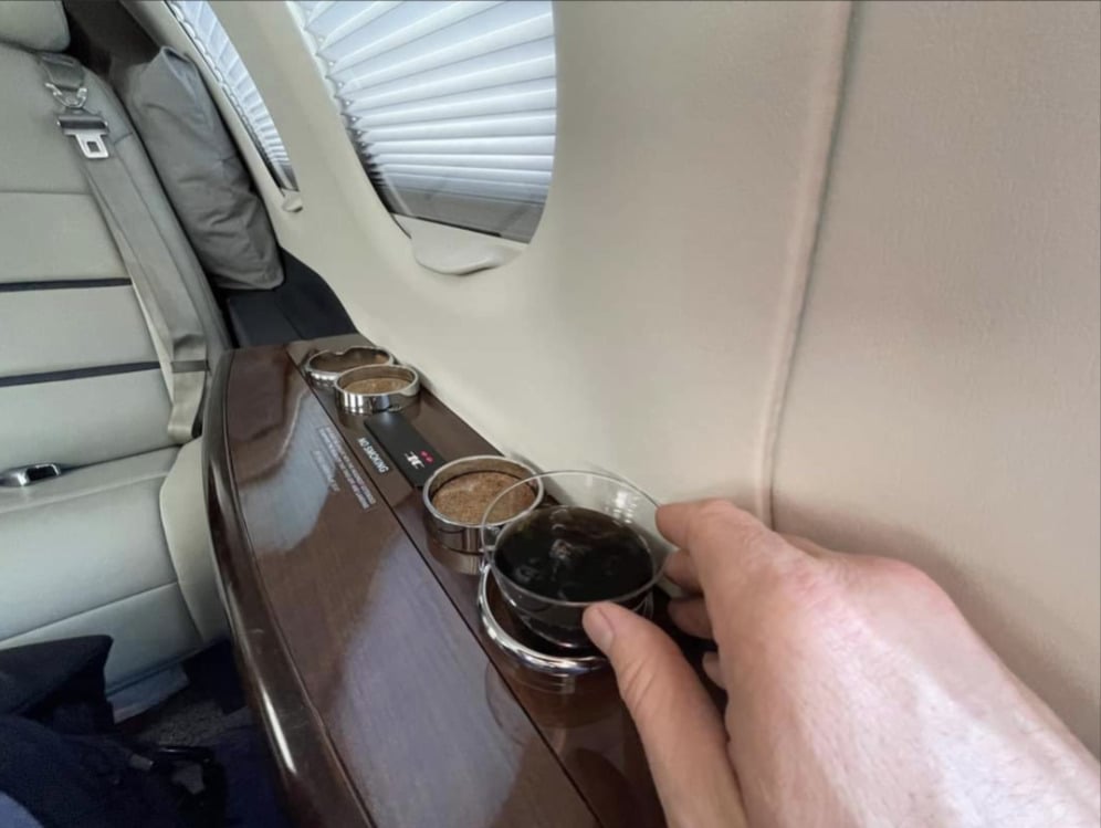 A hand holds a glass of dark soda on a polished wooden tray inside a private jet, with drink holders and window shades visible.