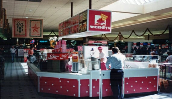 A man stands at a retro Wendy&rsquo;s counter in a mall food court, decorated with holiday garlands and banners. A worker in a pink uniform attends the counter under a red Wendy&rsquo;s sign.