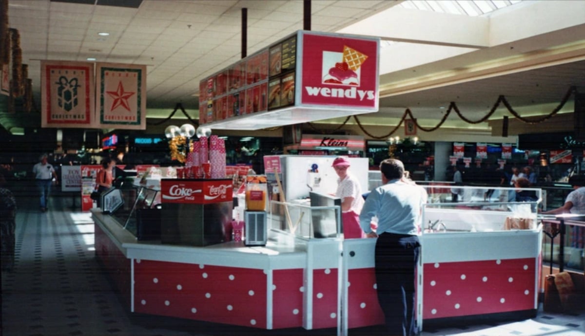 A man stands at a retro Wendy’s counter in a mall food court, decorated with holiday garlands and banners. A worker in a pink uniform attends the counter under a red Wendy’s sign.