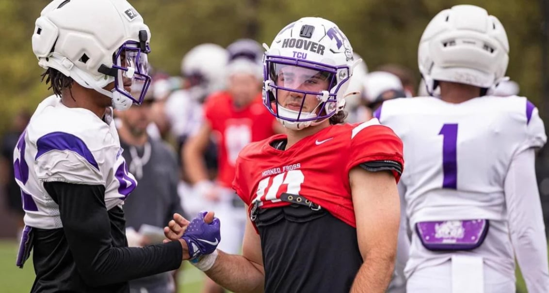 Two American football players, one in a red jersey and helmet and the other in a white and purple uniform, shake hands on a field. Other players in white and purple jerseys are visible in the background.