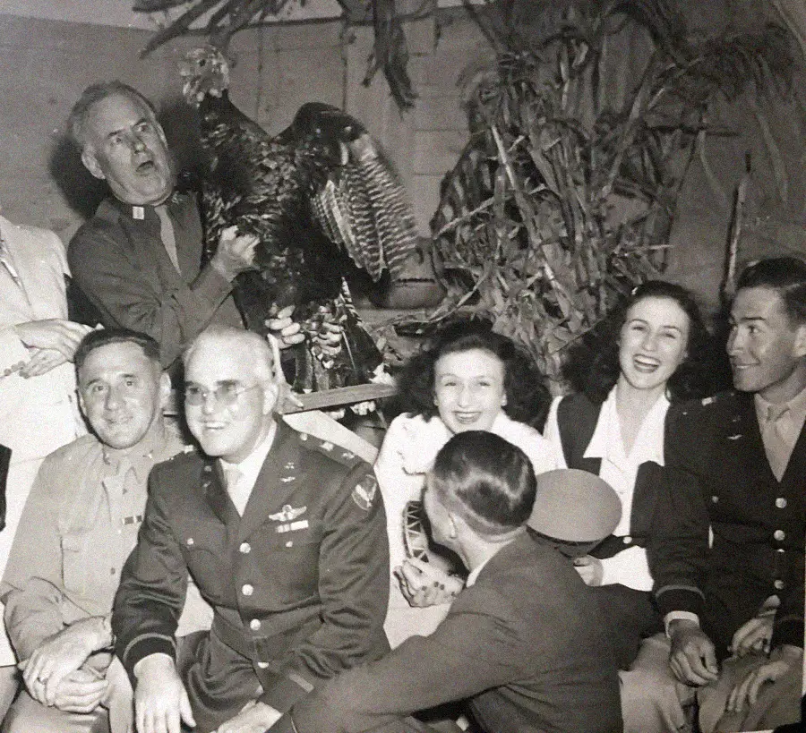 A group of people, some in military uniforms, sit and laugh together indoors. One man stands behind them, holding a large turkey. Dried cornstalks decorate the background.