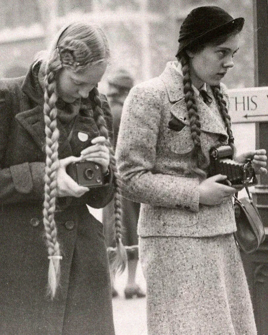 Two young women with long braids and coats stand side by side, each holding a vintage camera. One looks down at her camera while the other gazes ahead. The photo is black and white, suggesting a historical setting.
