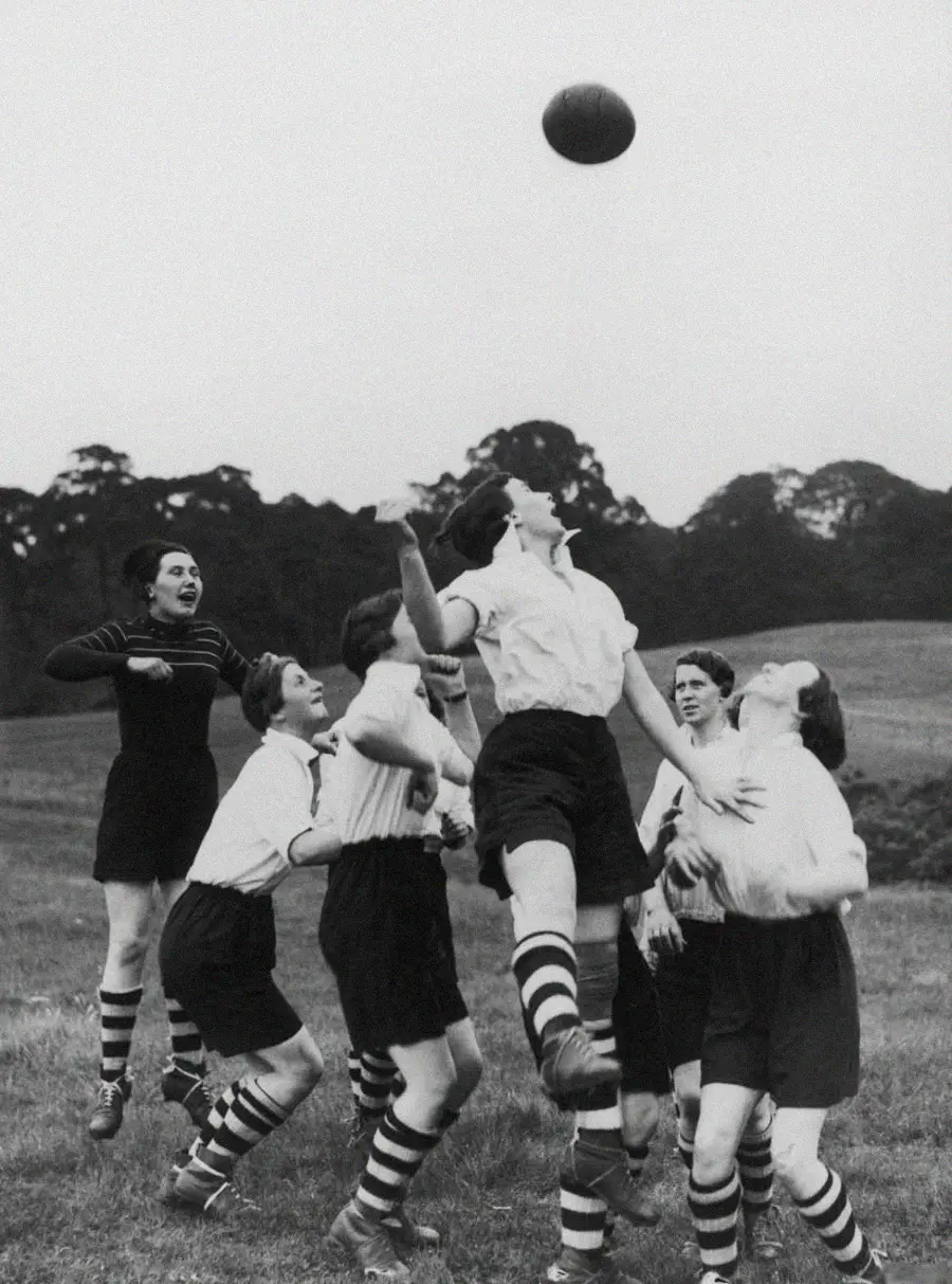 A group of women in vintage sportswear jump to reach a soccer ball in mid-air during a game on a grassy field, with trees visible in the background.