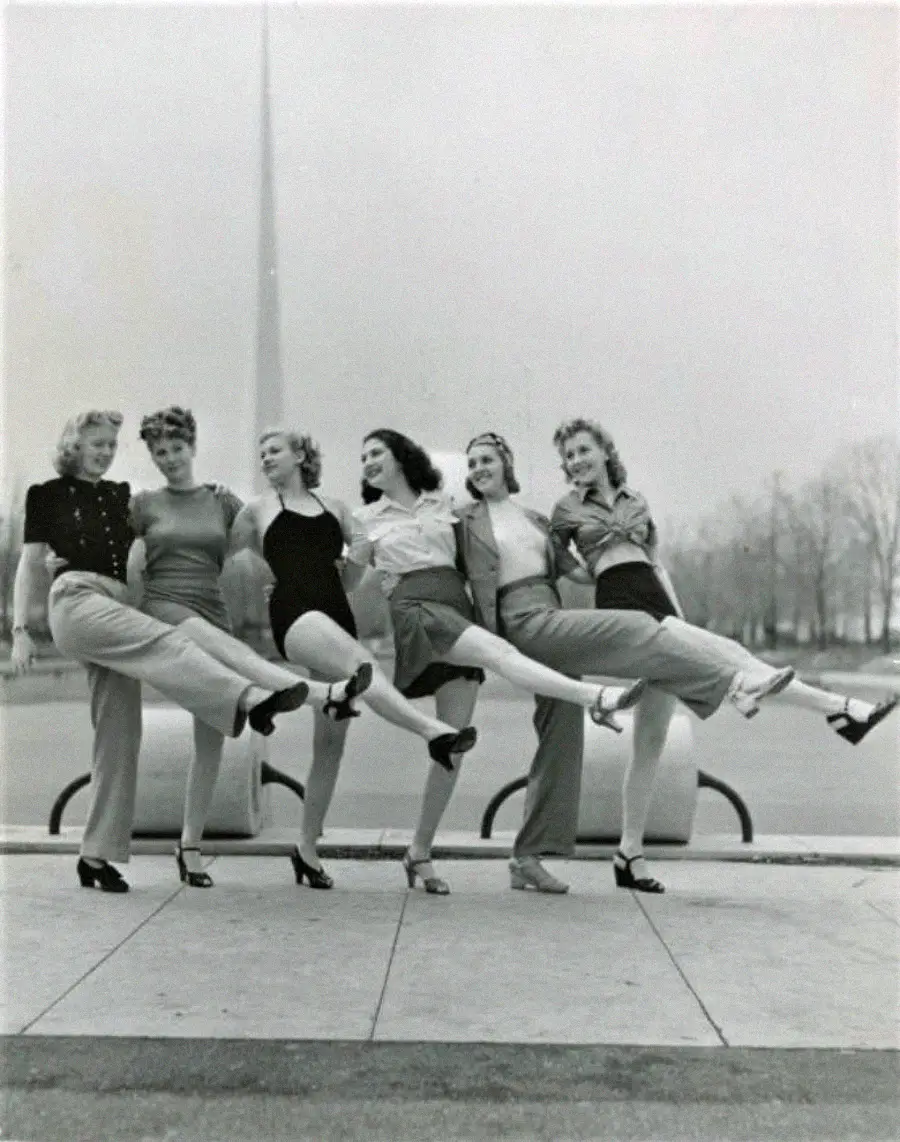 Six women pose outdoors in a line, kicking one leg up in unison and smiling. Leafless trees and a tall, thin monument or spire are visible in the background. The photo appears to be from the mid-20th century.