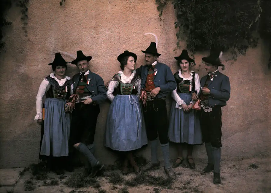 Six people in traditional European folk costumes stand against a textured wall. The group includes men and women wearing hats, vests, blouses, and aprons, with some greenery visible in the upper right corner.