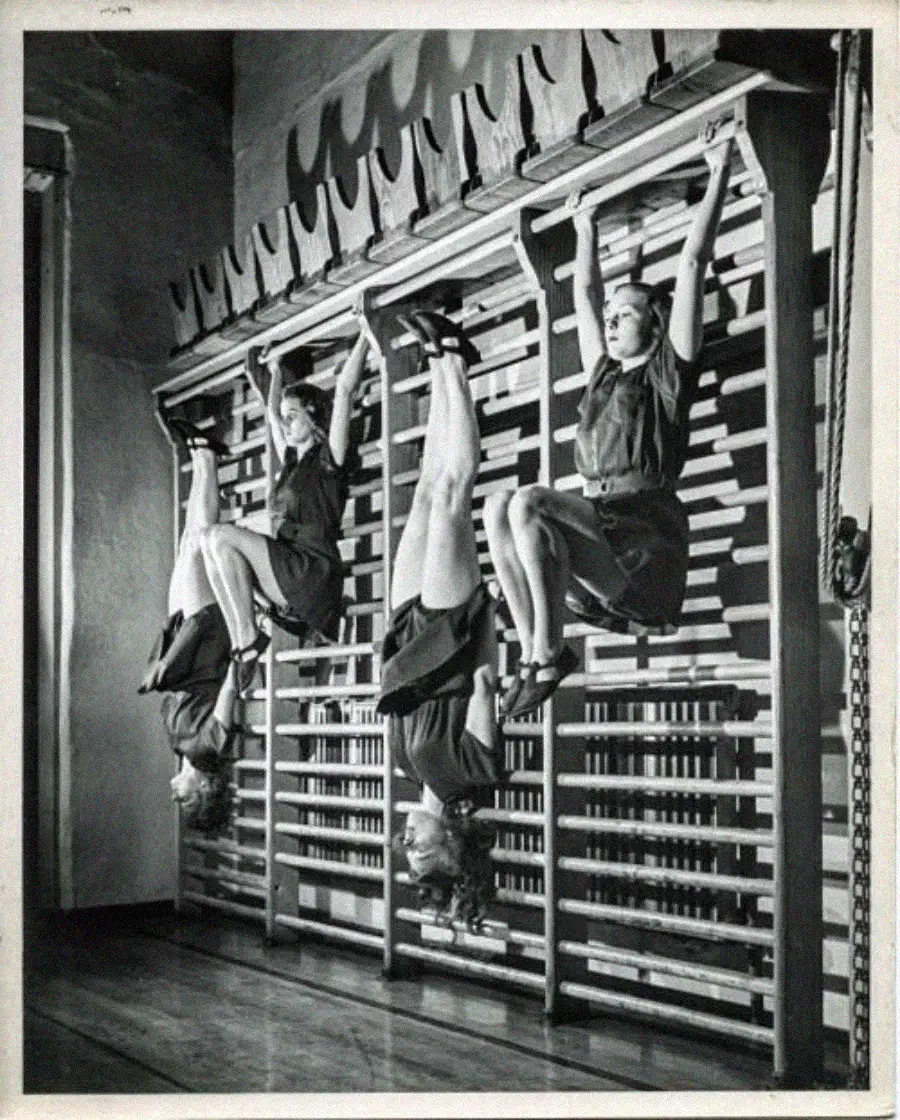 Three young women in dresses hang upside down from gymnastic wall bars in a vintage setting, gripping the bars with their legs and holding their arms out, performing a group exercise or gymnastic routine.