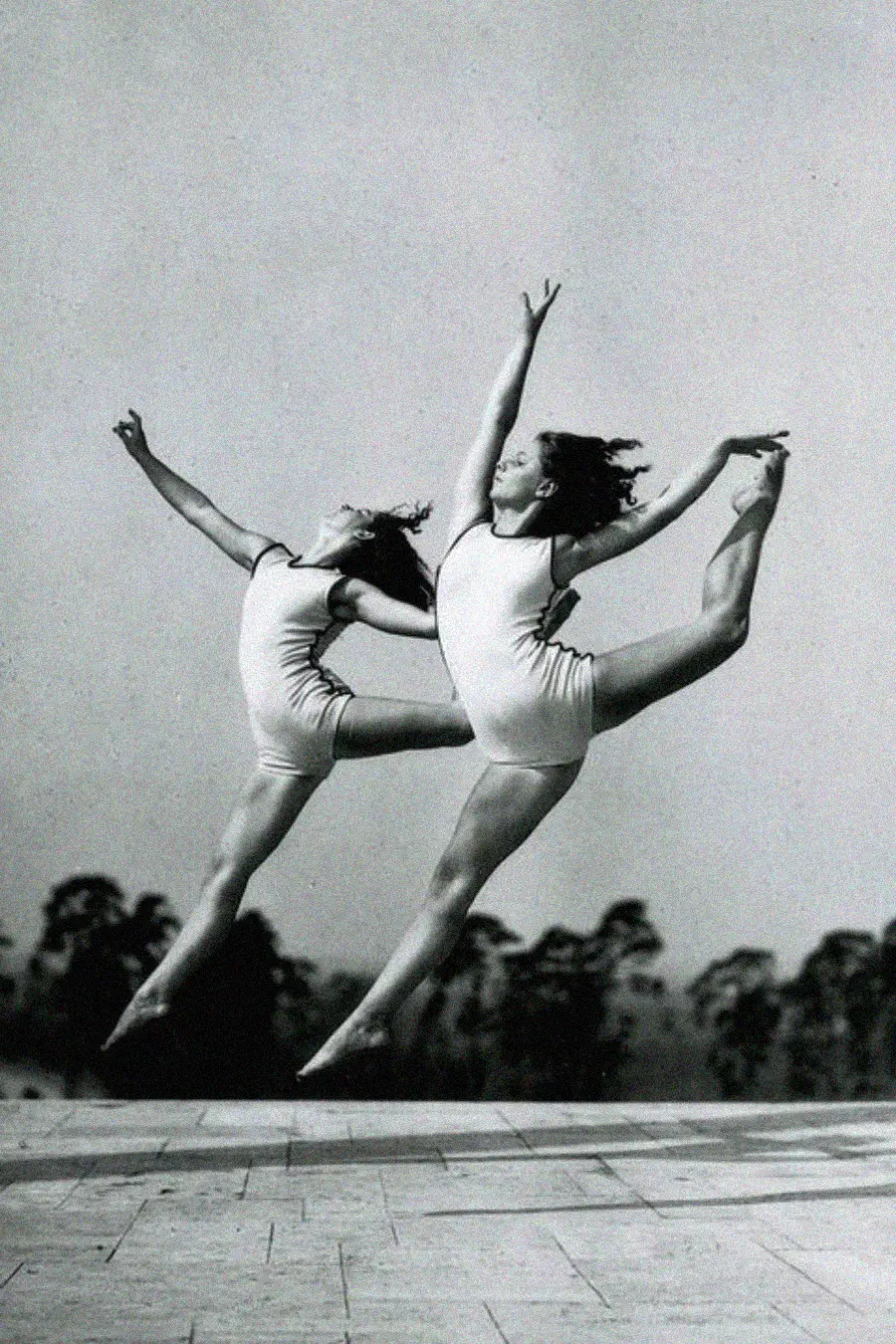 Two women in swimsuits leap gracefully in mid-air with outstretched arms and legs, performing synchronized dance or gymnastics outdoors against a blurred background of trees and sky.