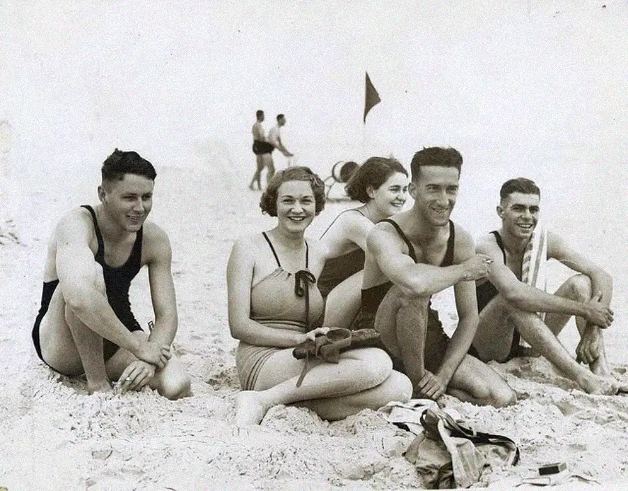 A group of five young adults in vintage swimsuits sit and smile on a sandy beach, with a flag and a few people in the background. The photo is black and white, capturing a cheerful, relaxed moment.