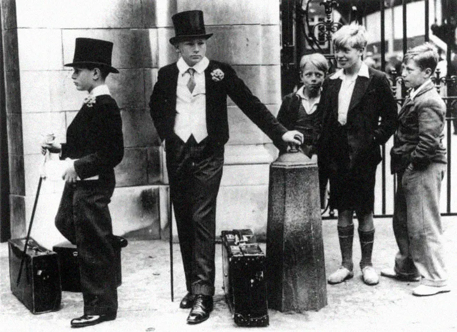 Five boys stand on a street; two wear formal suits with top hats and boutonnieres, while three others are dressed in casual, worn clothing and look on with curiosity.