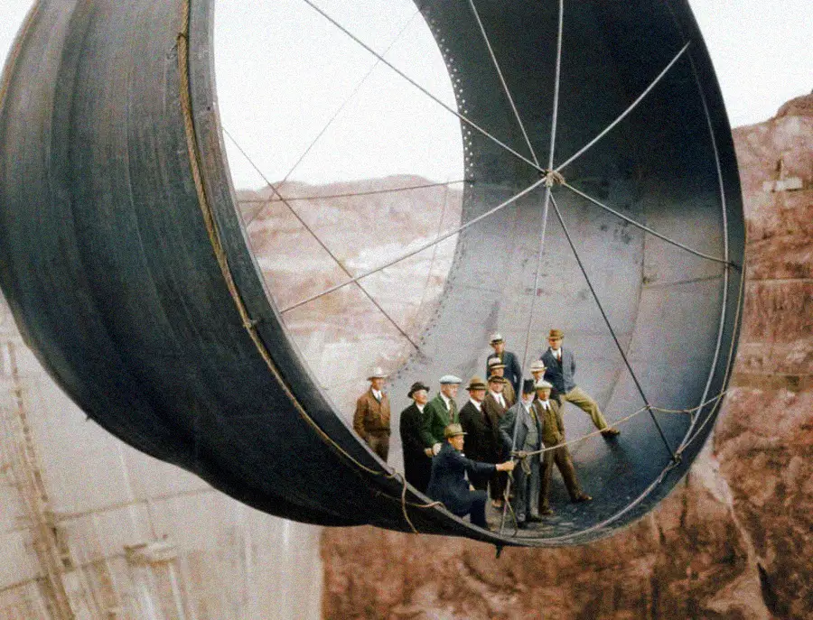 A group of men in suits and hard hats stand inside a large steel pipe segment suspended high above a rocky landscape, likely during the construction of a major infrastructure project.