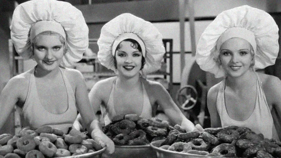 Three women in sleeveless outfits and large chef hats smile while holding trays piled high with donuts, in a black-and-white setting that appears to be a bakery or kitchen.