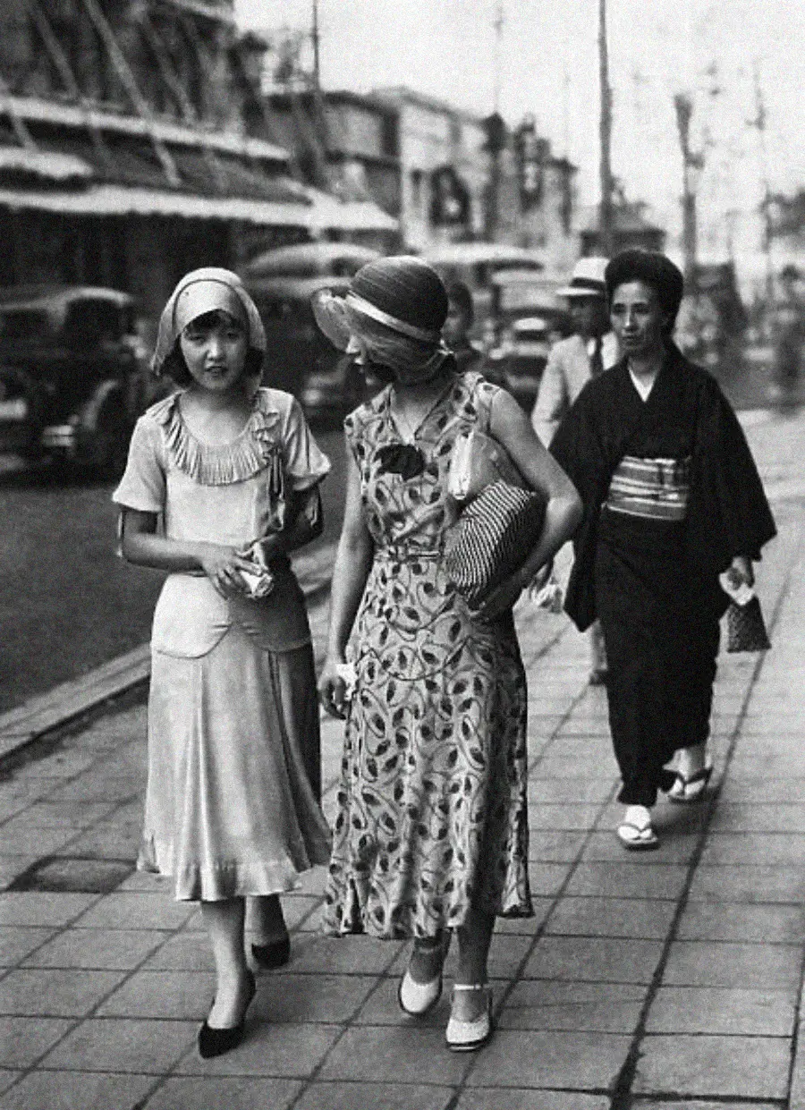 Two women in 1920s Western-style dresses and hats walk on a city sidewalk, talking. A woman in a traditional kimono walks behind them. Vintage cars and buildings line the street in the background.