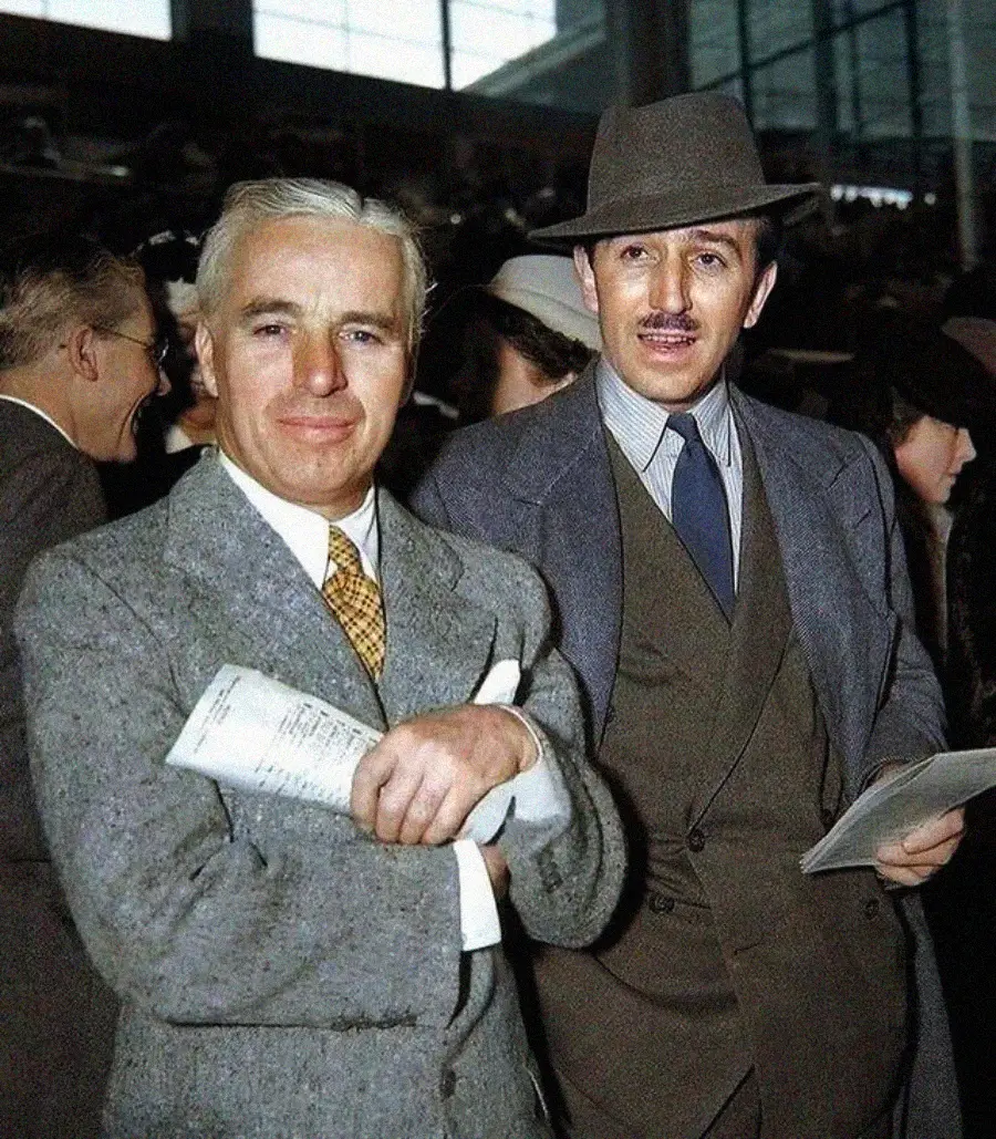 Two men in suits and ties stand together indoors among a crowd. The man on the left has gray hair and holds papers; the man on the right wears a hat and mustache. Both are smiling.