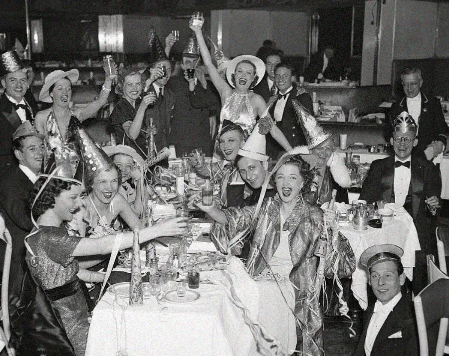 A black-and-white photo of people at a festive party wearing hats and costumes, raising glasses in celebration around decorated tables, smiling and cheering, with a lively, joyful atmosphere.