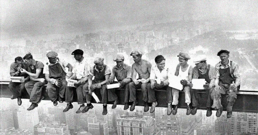 Eleven construction workers sit on a steel beam high above New York City, eating lunch and talking. Skyscrapers and a hazy city skyline are visible below them. The iconic photo is in black and white.