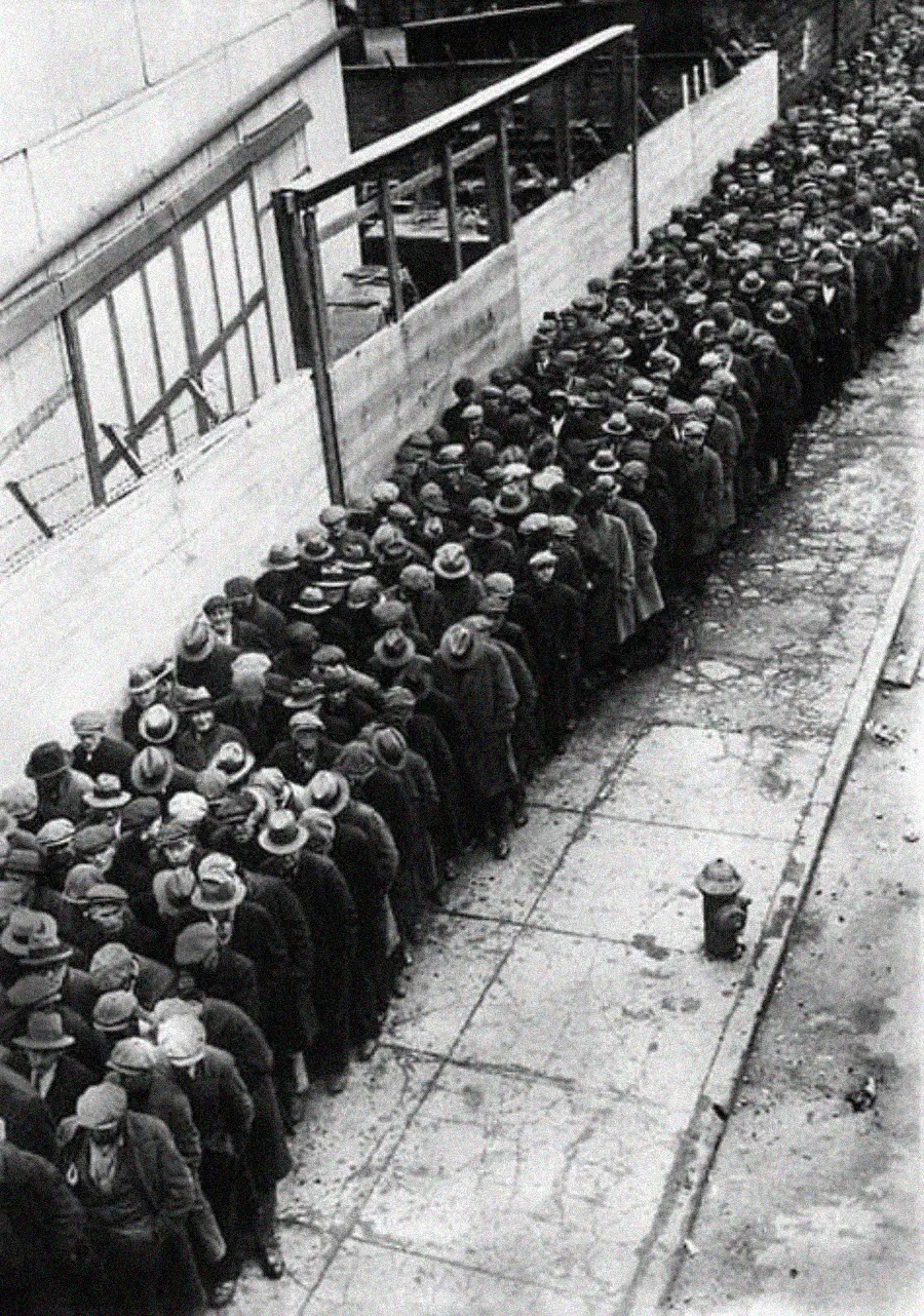 A long line of people wearing coats and hats stands on a city sidewalk next to a construction fence, viewed from above. The street looks worn and a fire hydrant is visible in the foreground.