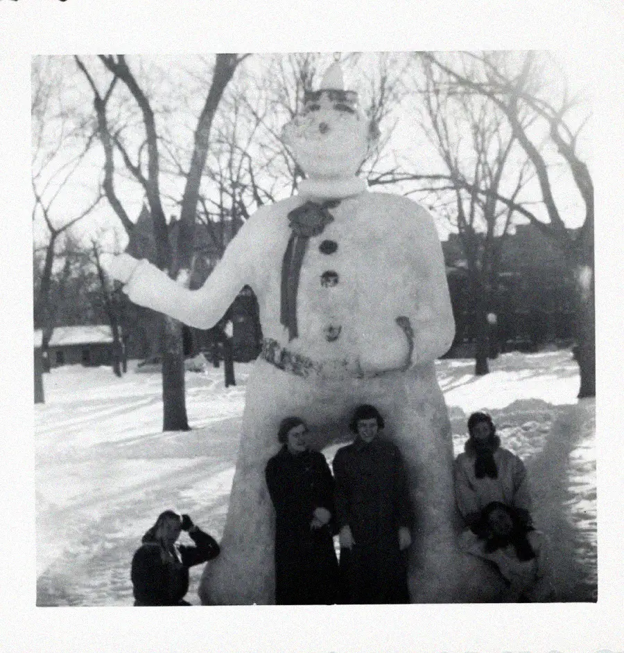 Four people pose in front of a large snowman statue in a snowy park. The snowman is decorated with buttons, a scarf, a belt, and a crown. Leafless trees and buildings are visible in the background.