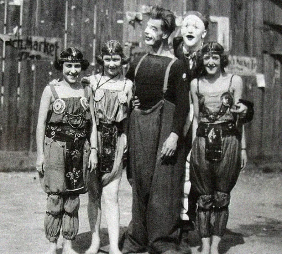 Four young women in elaborate costumes stand beside two clowns wearing face paint and baggy clothes, posing together and smiling in front of a wooden building. The scene appears to be from an old circus or carnival.