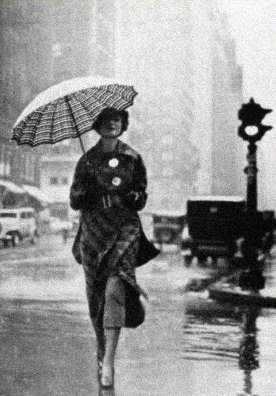 A woman walks on a rainy city street holding a striped umbrella. She wears a long coat and heels. Vintage cars and buildings are visible in the background, with wet pavement reflecting the scene.