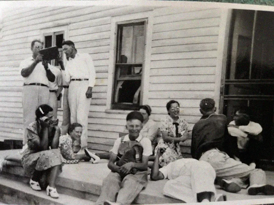 A group of people, including men, women, and children, sit and stand on the steps of a wooden building. Some are laughing, talking, or relaxing, creating a casual and joyful atmosphere.