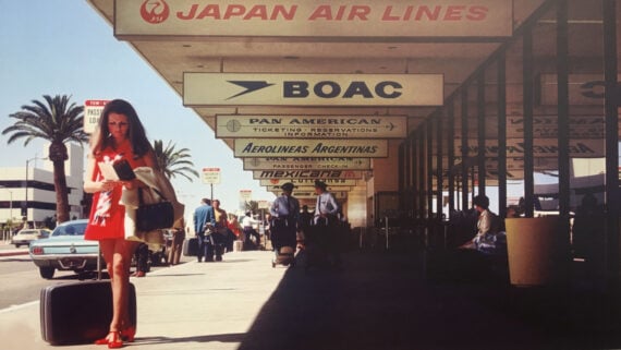A woman in a red dress and heels stands with a suitcase outside an airport terminal lined with retro airline signs, including Japan Air Lines, BOAC, Pan American, and Mexicana. Palm trees and cars are in the background.