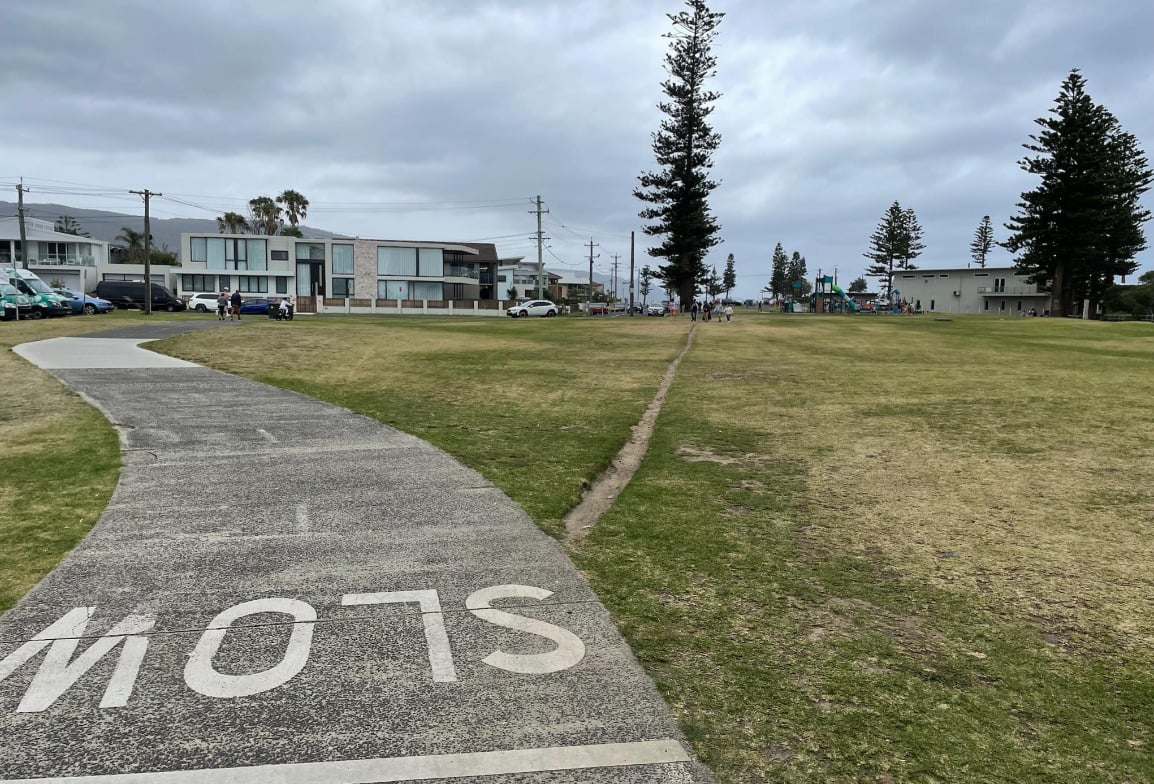 A paved path reading “SLOW” curves through a grassy park with modern houses on the left and a playground in the distance. Tall trees and cloudy skies complete the scene.