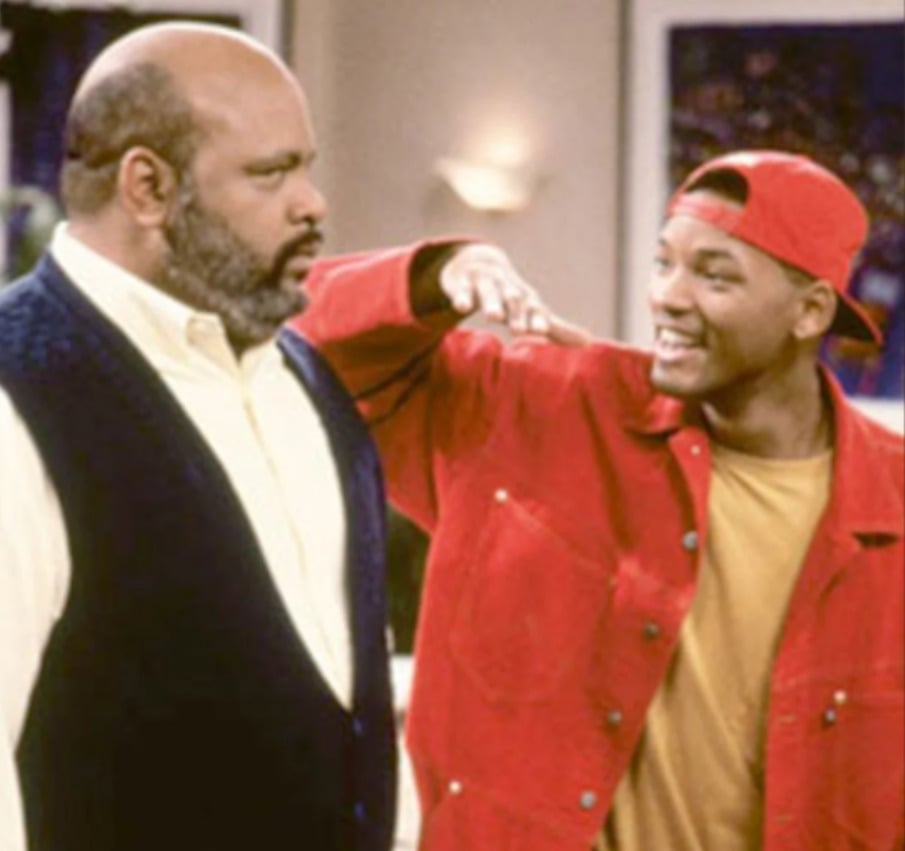 A young man in a red jacket and backwards cap playfully gestures towards an older man in a white shirt and dark vest, who looks at him seriously. They appear to be indoors, possibly in a living room.