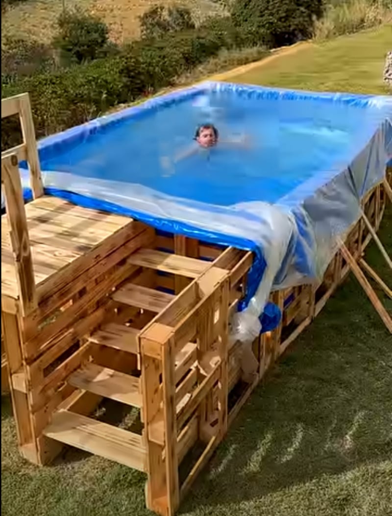 A person is swimming in a rectangular above-ground pool made with a wooden pallet frame and lined with blue plastic, set on grass with wooden steps leading up to it.