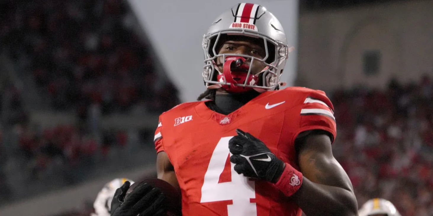A football player in a red Ohio State jersey with the number 4 and a silver helmet celebrates on the field while holding a football, with a blurred stadium crowd in the background.