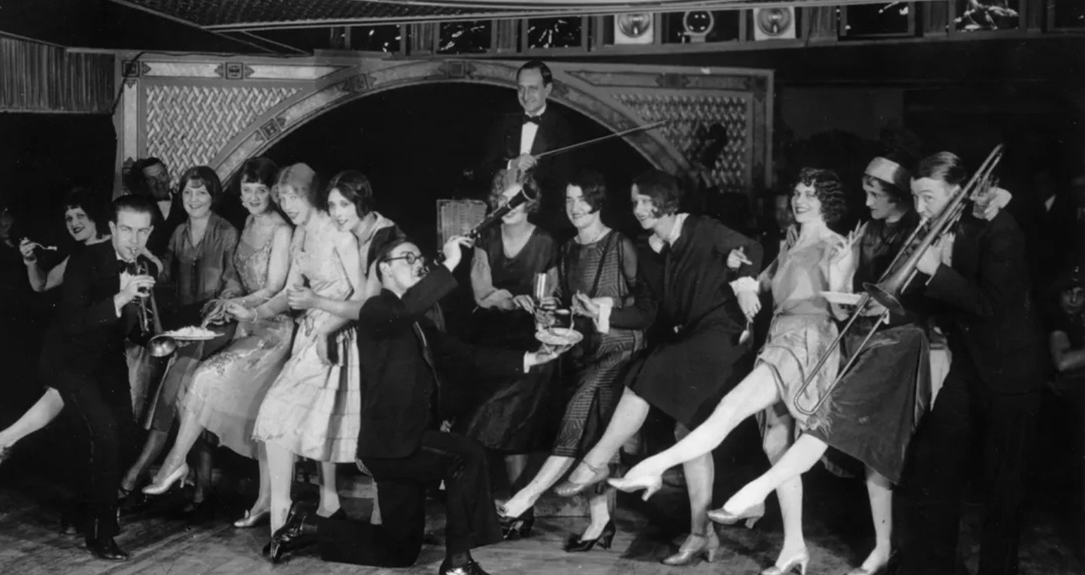 A lively black-and-white scene of men and women in 1920s attire dancing and kicking their legs, with musicians playing violin and saxophone beside them. The group appears joyful and energetic in a nightclub setting.