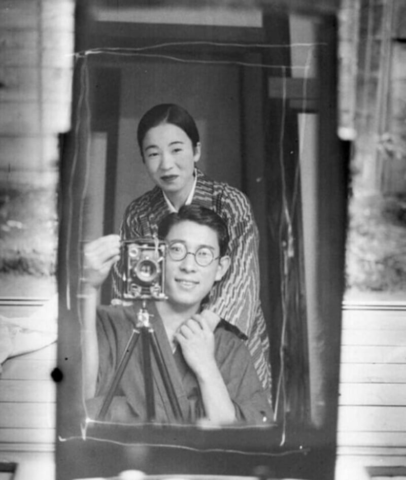 A black and white photo shows a man in glasses taking a mirror selfie with a vintage camera, while a woman in traditional clothing stands behind him, both looking into the mirror and smiling gently.
