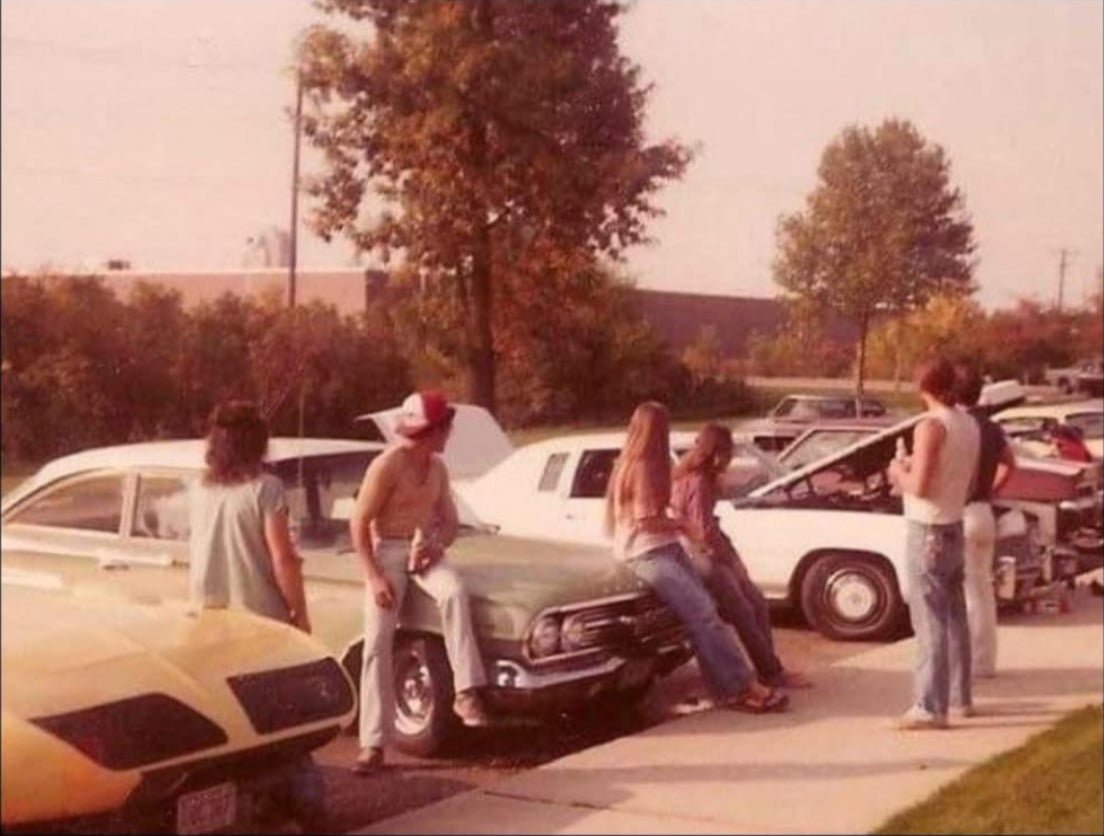 Four people in 1970s-style clothing hang out around classic cars in a parking lot, two sitting on a green car’s hood while others stand nearby. Trees and more cars are visible in the background on a sunny day.