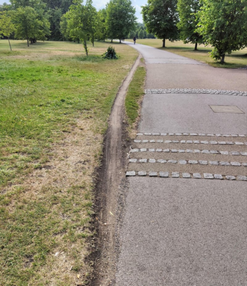 A worn dirt path runs alongside a paved walkway in a park with green grass, scattered trees, and a person walking in the distance.