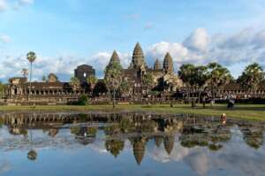 Angkor Wat temple in Cambodia is reflected in a pond, surrounded by palm trees, grass, and visitors under a blue sky with scattered clouds.