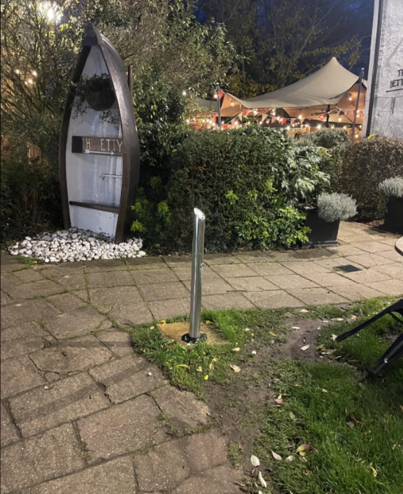 A small garden area with a boat-shaped decorative sign that reads "HETTY," surrounded by white stones and bushes. A metal pole stands on a paved path, with a tent and string lights visible in the background.