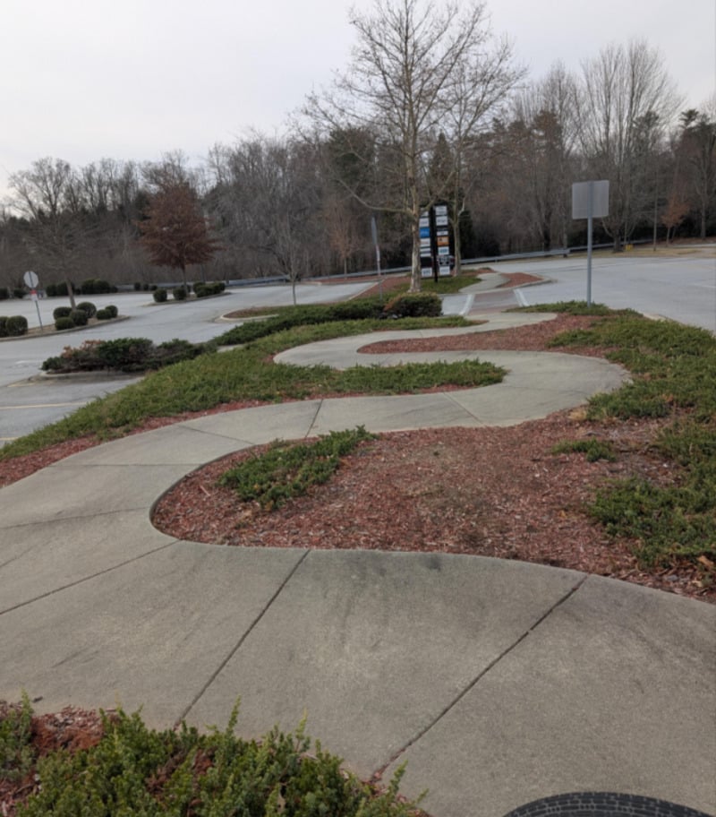 A wavy, curving concrete sidewalk winds through landscaped greenery and mulch in an empty parking lot with leafless trees and cloudy skies in the background.