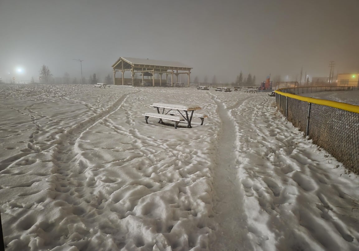 A snowy park at night with many footprints and a picnic table in the center. A pavilion stands in the background, lights glow in the distance, and a fence runs along the right side of the image.